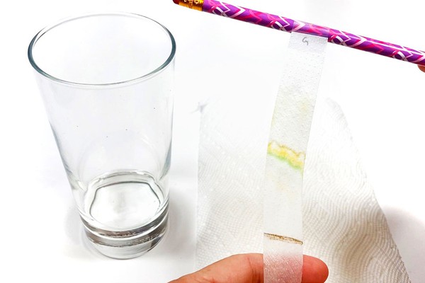 A tall glass with some liquid in it is standing on a table. Next to the glass a person is holding a paper strip. The paper strip is wet and shows orange, yellow, and green lines across its length.