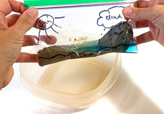 Hands holding a small plastic bag that contains some colored water, sand, and a small rock.