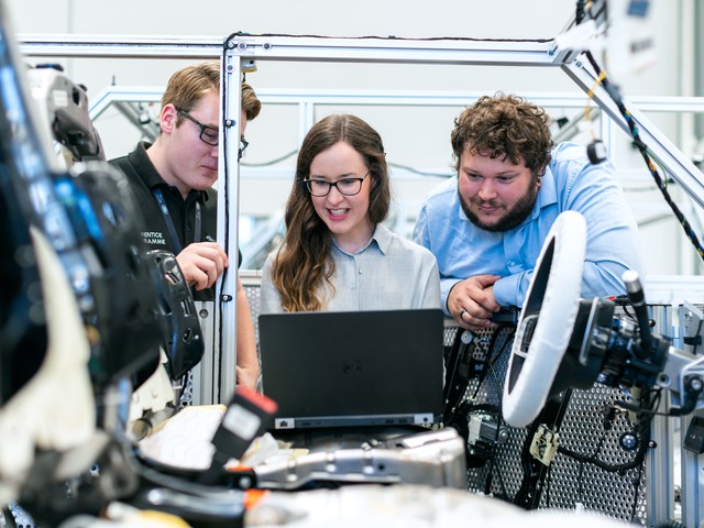 three people looking at data on a laptop in a car under construction