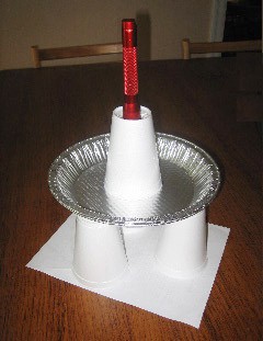 Three upside down cups hold a pie pan with a fourth cup placed on top that has a flashlight protruding from the bottom