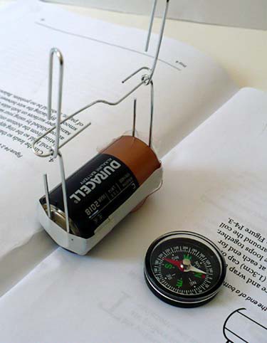 A paperclip is suspended between two paperclips that are taped to the terminals of a C cell battery next to a compass