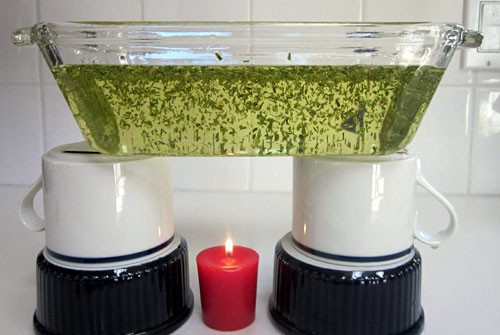 A lit candle sits under a glass dish filled with oil and dried thyme that is held up by two upside down cups