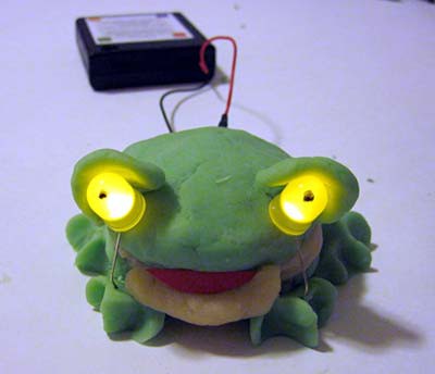 A frog is made of model clay and has two LEDs for eyes that are powered by a battery pack