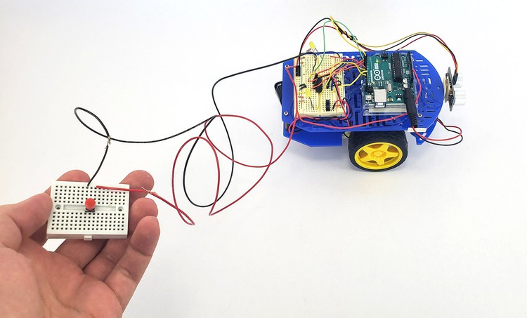  A hand holds a mini breadboard remote wired to a robot vehicle.