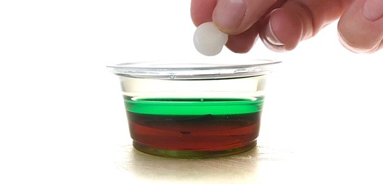 A hand is holding a small piece of candle wax next to a mini cup filled with dark corn syrup, green water, and vegetable oil.