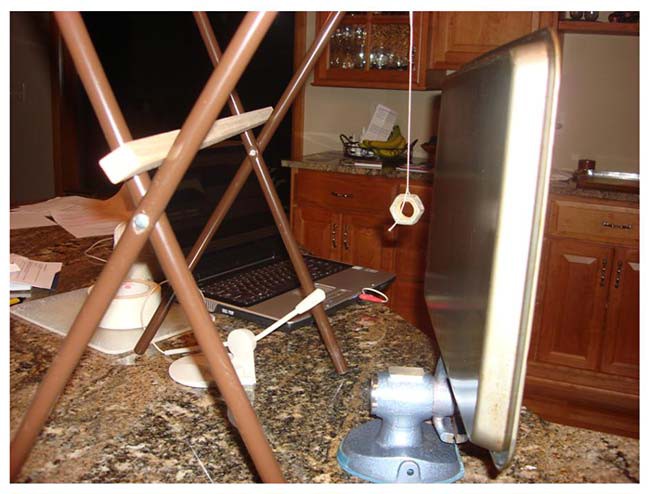 A vise holds a baking tray vertically next to a nut that is suspended from a string