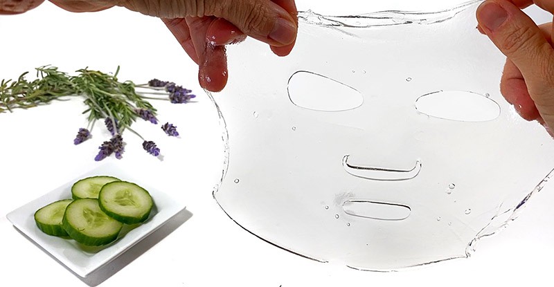 Two hands holding a collagen hydrogel mask next to a small plate with cucumbers and a bunch of lavender flowers.  