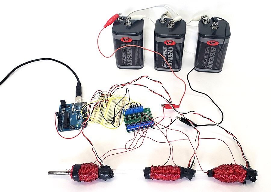 An Arduino, a circuit board, three lantern batteries, and a clear plastic tube with three coils of wire around it. 