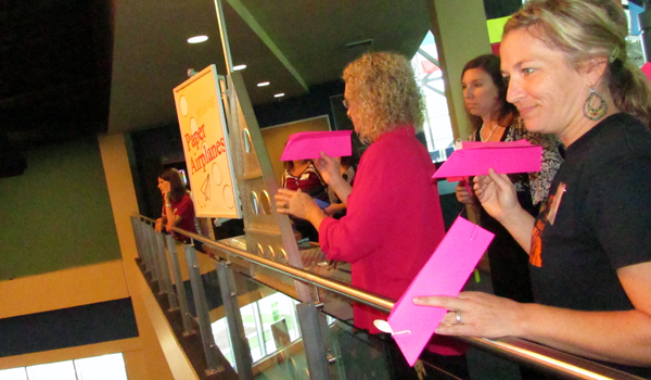 Two women prepare to throw pink paper airplanes over the rail of a balcony