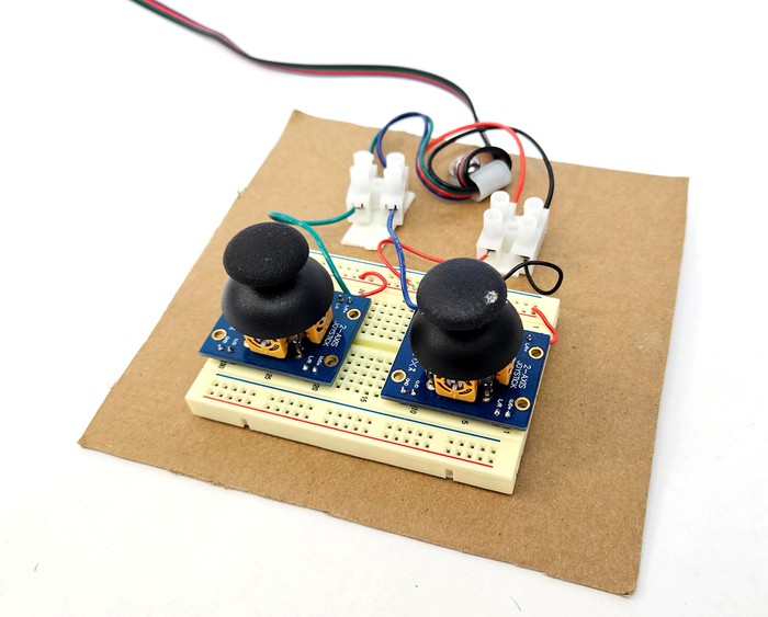 A piece of cardboard with a breadboard, two joysticks, and wire clips mounted on it 