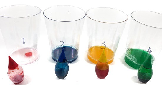 Four cups with different colored solutions. The respective food color vials are placed in front of each cup.
