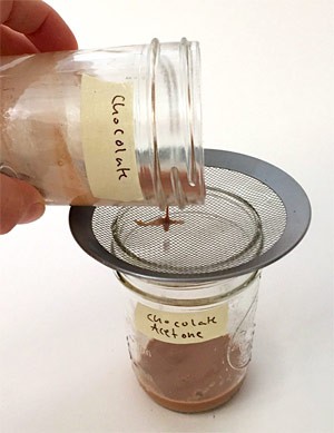 Crushed chocolate and acetone are poured through a mesh strainer into a glass jar