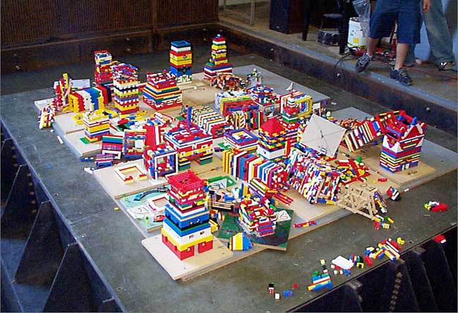 A table is filled with LEGO buildings, some of which have collapsed or fallen over