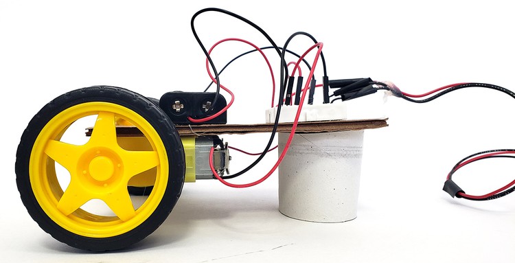 Side view of cardboard robot body, with battery pack and breadboard on top, and motors with wheels on the bottom. A short piece of cardboard tube keeps the body level since there are only two wheels.  