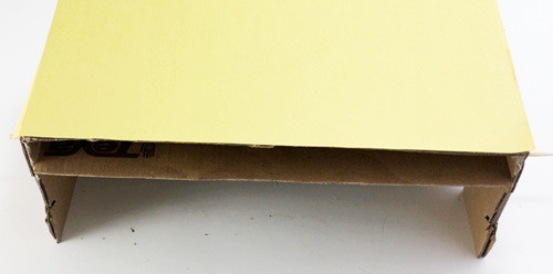 A shoebox placed upside down with the front panel removed. A piece of cardboard is placed in the box, parallel to the top of the box. 