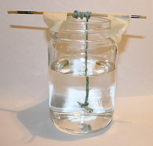 A screw tied to a string is suspended in a glass jar filled with sugar water
