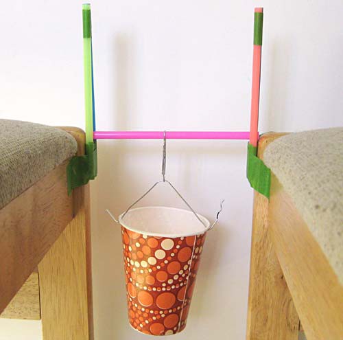 A paper cup is suspended from the center of a straw that rests on two tower bridges made of straws
