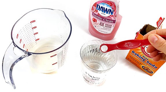 A measuring cup, dish soap, and baking soda are standing next to a cup filled with water. 1/8th teaspoon of baking soda is added to the cup.