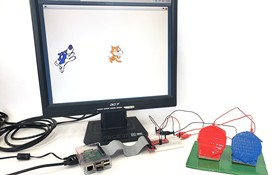 A raspberry pi, computer monitor showing Scratch, and adaptive game controller with two large buttons 