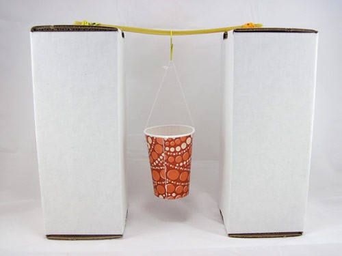 A paper cup hangs from the center of a birdge made of dry spaghetti