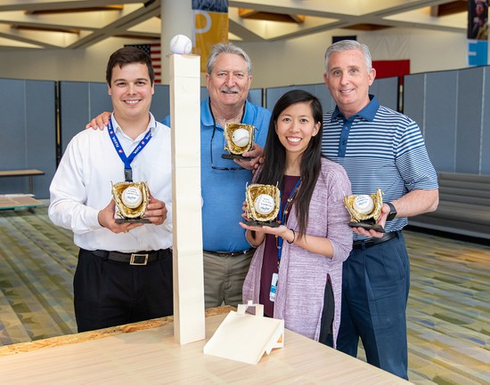 Three male and one female engineers standing around a three-foot-tall paper tower balancing a baseball on top.