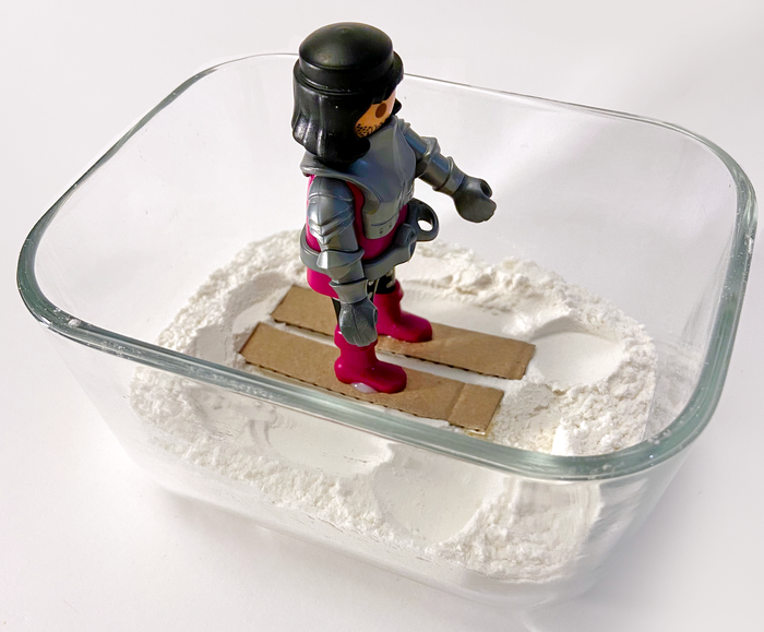 Toy figure with cardboard skis standing in a small container of flour pretend snow
