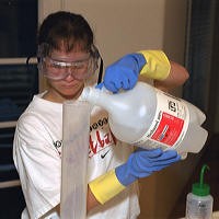 A student pours a bottle of liquid into a large graduated cylinder