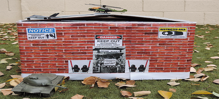 A toy tank, jet and helicopter surround a model building