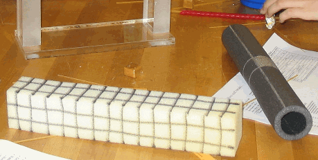 A rectangular prism made of foam with grid lines drawn on each side next to a foam tube