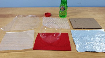 A soap bubble rests on a square sheet of red felt next to square sheets of other materials