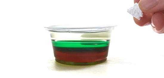 A hand is holding a small piece of Styrofoam next to a mini cup filled with dark corn syrup, green water, and vegetable oil.