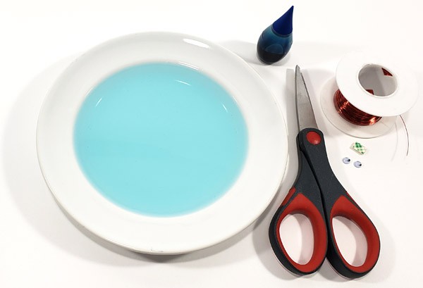 A plate filled with water, blue dye, scissors, a spool of copper wire, double sided tape and googly eyes