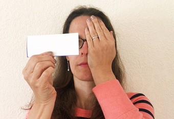 A person covering her left eye with her left hand. In her right hand she holds a paper card close to her face at eye level.