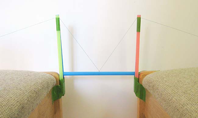 A straw rests length-wise between two tower bridges made of straw and has its center supported by two tensioned threads