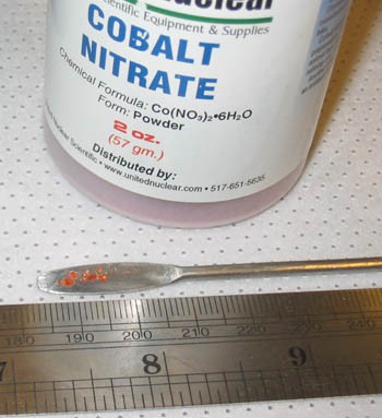 Eight grains of orange cobalt nitrate on a metal scoop next to a ruler