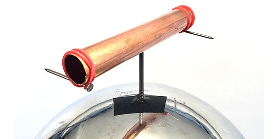 A copper pipe balanced on top of a nail, which is taped to the top of a Van de Graaff generator. The pipe has two additional nails sticking out from its ends, held in place by rubber bands, and pointed in opposite directions. 