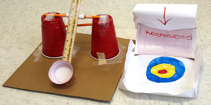 A ball launcher made from a ruler, pencils, tape and plastic cups next to a paper target