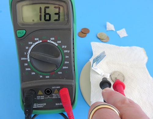 Two leads of a multimeter touch an aluminum foil strip and a stack of a penny, paper towel, and nickel to measure the voltage