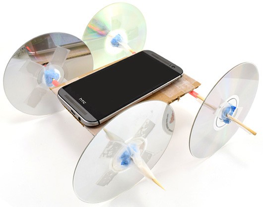 A smartphone rests on a vehicle with CD's as wheels