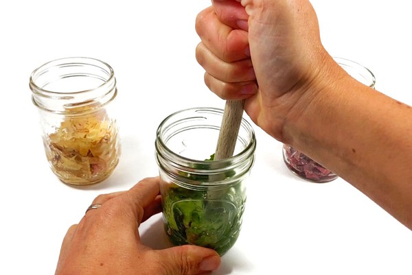 A mason jar with cut green leaves standing on a table. A person is crushing the leaves with the blunt end of a wooden spoon.