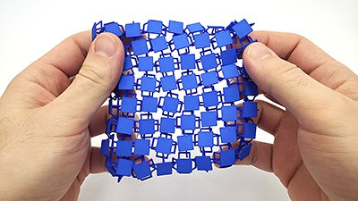 Two hands hold a piece of 3D-printed fabric consisting of a grid of small squares connected by thin loops 