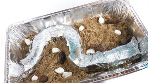 An aluminum pan that has a river model made from aluminum foil inside. Water is poured from a bottle into the aluminum riverbed  