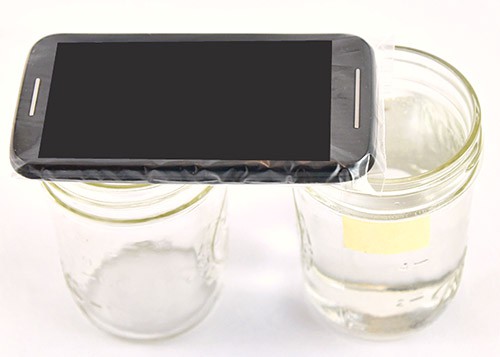 A smartphone in a plastic bag is placed on the rim of a glass jar so the microphone is above the rim of a second glass jar
