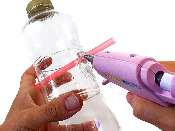 Hands holding a glue gun that puts glue around a straw that is inserted through the sides of a plastic bottle.