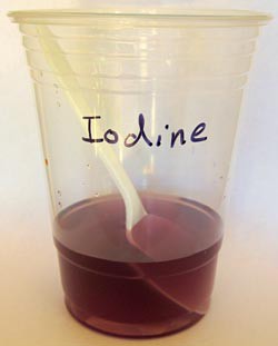 A violet colored solution and plastic spoon sit in a plastic cup labeled iodine