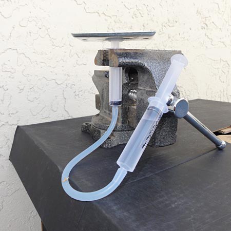 A syringe is held upright in a vise while being connected at the tip to another syringe through a plastic tube