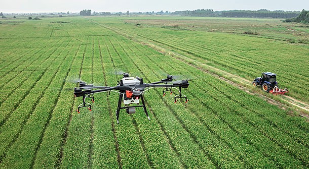 Drone flying above field with rows of crops and a tractor.