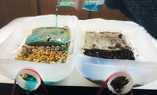 Two mini rain garden models, one with pebbles and the other with soil, next to each other. Blue water is being poured on one of the gardens.