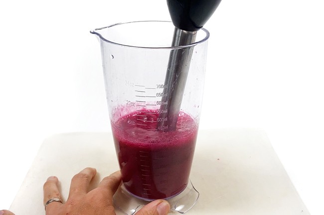An immersion blender is mixing a red solution in a measuring cup. 