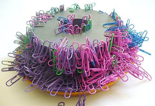 A magnet in the shape of a ring sits on a pile of multi-colored paperclips